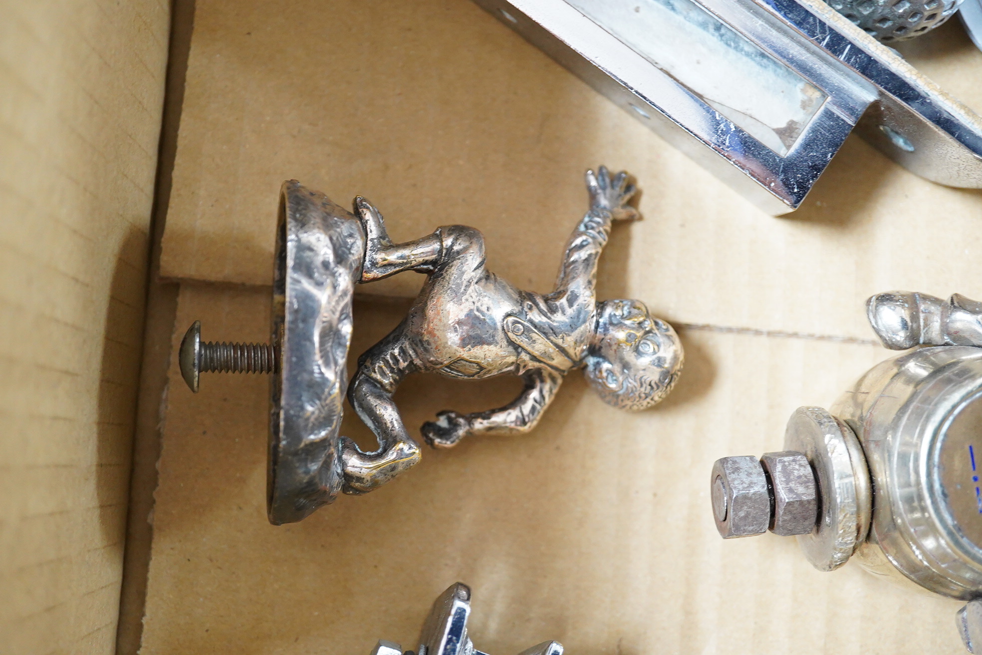 Seven chrome and brass car mascots, including a Mercedes-Benz logo, a figure of a blacksmith, a caricature figure on a beer barrel, a golf ball in mid flight, etc. together with a chromed housing unit for an electric lam